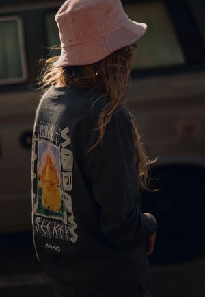 Czarna koszula z długim rękawem z graficznym wzorem przedstawiającym kolorową górę i napisem "SUNSET SEEKER". Miękka faktura, noszona z różowym kapeluszem typu bucket.
