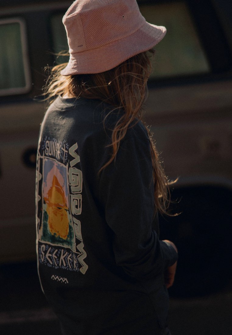 Czarna koszula z długim rękawem z graficznym wzorem przedstawiającym kolorową górę i napisem "SUNSET SEEKER". Miękka faktura, noszona z różowym kapeluszem typu bucket.