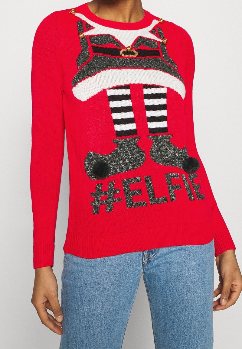 Pull rouge avec motif jambes et bottes d'elfe, détails boutons dorés et texte noir hashtag "ELFIE", porté avec un jean bleu clair.