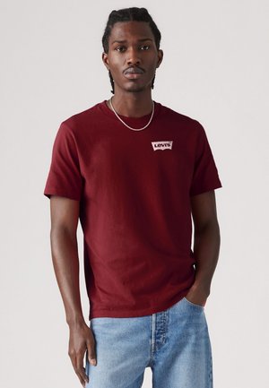 Junger Mann mit geflochtenem Haar, der ein burgunderfarbenes Levi's T-Shirt und hellblaue Jeans trägt, steht vor einem schlichten grauen Hintergrund.
