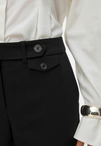 Primer plano de pantalones negro de cintura alta con detalle de doble botón, combinados con una blusa blanca que lleva una pulsera de brazalete dorado.