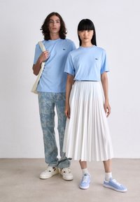 Zwei Models stehen nebeneinander und tragen hellblaue Lacoste-T-Shirts; einer in gemusterten Jeans und weißen Sneakers, der andere in einem weißen Plisseerock und blauen Sneakers.