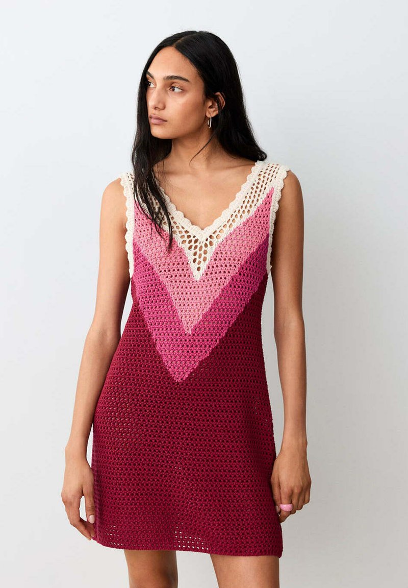 Femme aux longs cheveux foncés portant une robe sans manches en crochet aux nuances bordeaux et roses, regardant vers sa gauche sur un fond uni.