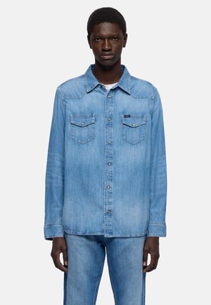 Giovane uomo che indossa una camicia di jeans azzurro chiaro con due tasche sul petto e jeans di jeans coordinati, in piedi davanti a uno sfondo semplice.