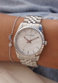 Silberner Edelstahluhren mit einer gewellten Lünette, Zifferblatt aus Perlmutt und roségoldenen Zeigern. Gliederarmbandstil. Kettenarmband mit Halbmond-Anhänger.