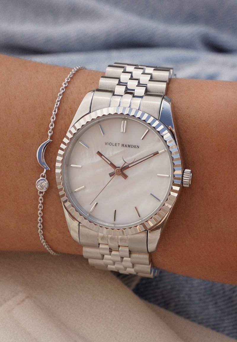 Silberner Edelstahluhren mit einer gewellten Lünette, Zifferblatt aus Perlmutt und roségoldenen Zeigern. Gliederarmbandstil. Kettenarmband mit Halbmond-Anhänger.