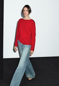 Ein Modell, das selbstbewusst geht und einen roten Pullover sowie weit geschnittene hellblaue Jeans trägt, vor einem minimalistischen Hintergrund in Weiß und Schwarz.