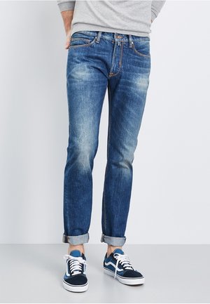 Mężczyzna ubrany w podwinięte niebieskie dżinsy i niebiesko-białe trampki, stojący na jednolitym jasnym tle.