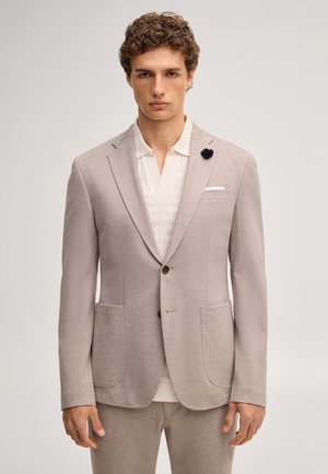 Man draagt een beige colbert met een zwarte reversspeld en een wit gestructureerd overhemd, staand tegen een effen lichte achtergrond.