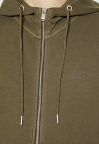 Sudadera con capucha verde olivo con cremallera, hecha de tela suave, con capucha forrada, cremallera plateada y un logo sutil en el pecho.