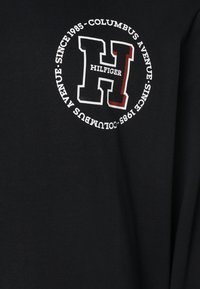 Czarna koszula z długim rękawem z wyraźnym okrągłym logo z białą i czerwoną literą "H", z napisem "HILFIGER" oraz "COLUMBUS AVENUE".