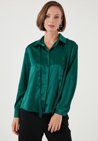 Satin emerald green blouse with a collared neckline, long sleeves, and front button closure. Features vertical pleats and a smooth texture.
