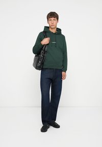 Sweatshirt à capuche verte avec cordons, jeans en denim foncé et sac fourre-tout noir. Le modèle porte des chaussures noires. Fond uni.