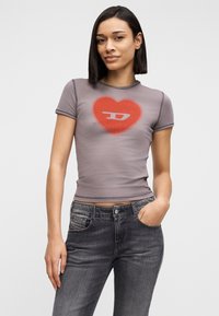 Γυναίκα με ίσια σκούρα μαλλιά, φοράει εφαρμοστό γκρι μπλουζάκι με κόκκινη καρδιά και λογότυπο, σε συνδυασμό με ξεβαμμένο μαύρο τζιν, με το χέρι στην τσέπη.