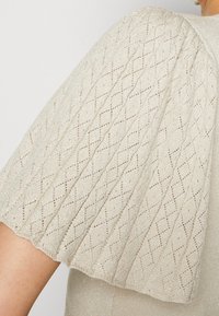 Manche courte avec texture en maille à motif losange sur tissu beige clair porté par une personne visible de l'épaule jusqu'au milieu du bras.