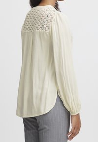 Blouse en crème clair avec des manches longues, en tissu texturé, et un panneau en dentelle dans le haut du dos. Fentes latérales pour faciliter le mouvement, silhouette semi-ajustée.