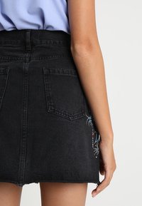 Svart denimkjol med fransad fåll och två bakfickor, prydd med färgglad spindelbroderi längs sidan.