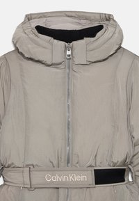 Világosszürke puffertömörítő, magas gallérral, elülső cipzárral és hímzett "Calvin Klein" logóval a derékrészen. Simább textúrájú.