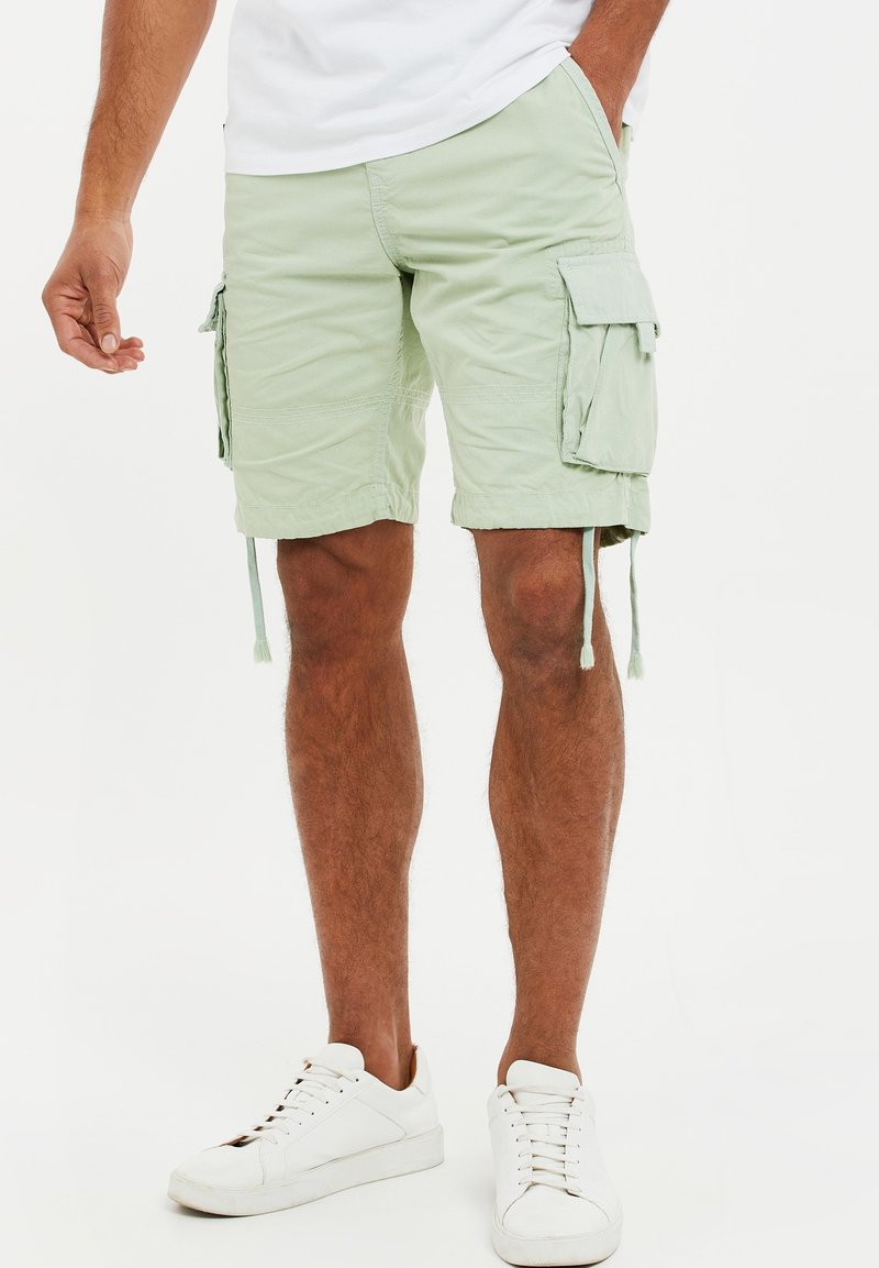 Homme portant un short cargo vert pâle avec des poches latérales et des baskets blanches, debout avec une main dans la poche.