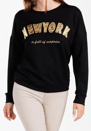 Woman wearing black sweatshirt with shiny gold "NEW YORK" text and smaller script "is full of surprise," paired with beige pants.