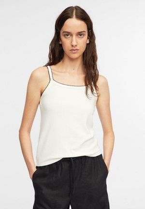 Giovane donna con capelli castani lunghi che indossa un top bianco con bordatura nera e pantaloni neri, con le mani nelle tasche, in piedi su sfondo bianco.