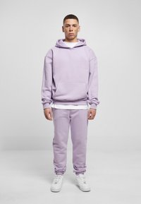 Felpa in pile lavanda con tasca frontale, abbinata a jogger coordinati. Presenta polsini elasticizzati e una maglietta bianca a strati sotto. Sneakers bianche.