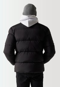 Chaqueta puffer negra con textura acolchada, vista desde atrás mostrando una sudadera con capucha blanca debajo y un gorro de punto gris arriba.