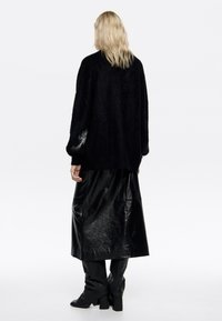 Oversize'owy czarny sweter z długimi rękawami i misternym piórowym zdobieniem na lewym ramieniu, zestawiony z błyszczącą czarną skórzaną spódnicą.