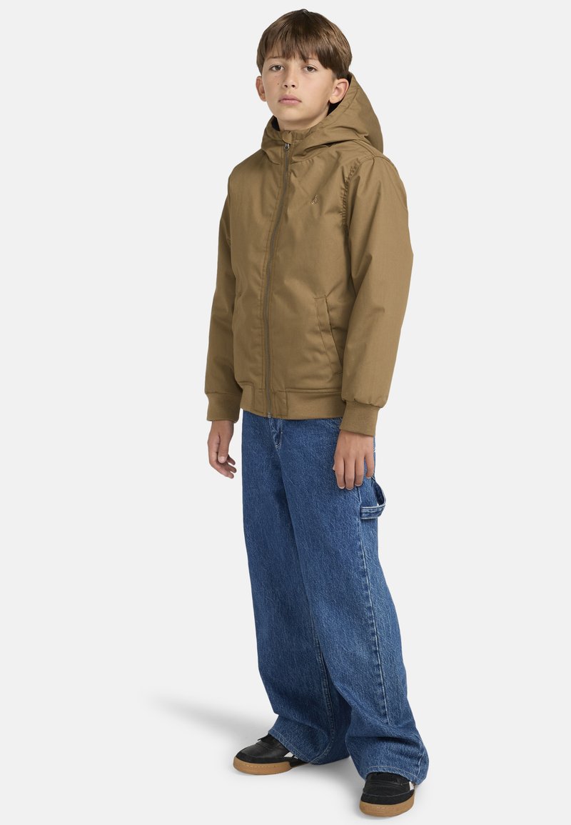 Brun zip-up jakke med hætte, med sidelommer og ribbede manchetter, parret med løse blå denimjeans og sorte sko.