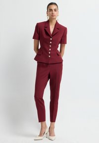 Bordeaux maatpak met blazer met korte mouwen, voorzien van gouden knopen en peplumdetail, gecombineerd met slim-fit broek en crèmekleurige hakken.