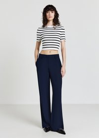 Weißes Crop-Top mit schwarzen horizontalen Streifen, kombiniert mit marineblauen weit geschnittenen Hosen und schwarzen spitzen Schuhen. Glatter Stoff, maßgeschneiderter Schnitt.