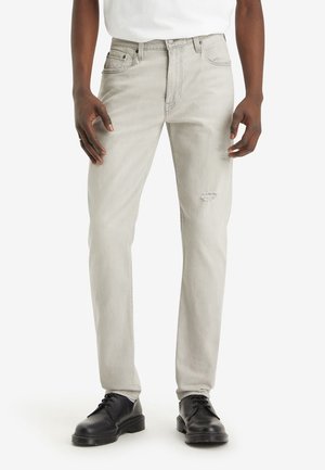 Person iført lysegrå slim-fit jeans og sorte snørebåndssko står mod en ensfarvet hvid baggrund.