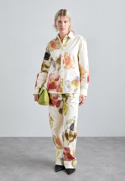 Completo di camicia e pantaloni stampati floreali in crema, caratterizzato da fiori vibranti rossi, gialli e neri. L'outfit è abbinato a una pochette verde.