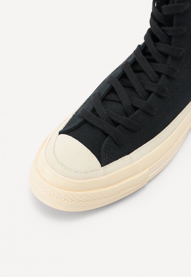 Zapatilla alta de lienzo negro con punta y suela de goma crema. Presenta cordones negros y costuras reforzadas a lo largo de las costuras.