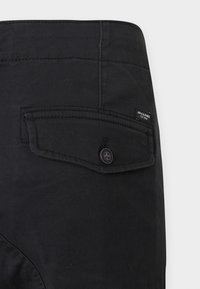 Pantalon en coton noir avec une poche arrière boutonnée. Présente des coutures discrètes et une petite étiquette de marque sur la ceinture.