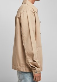 Beige bomullsjacka med avslappnad passform, långa ärmar, knäppning och en framficka. Sidovy visar en skräddarsydd struktur.