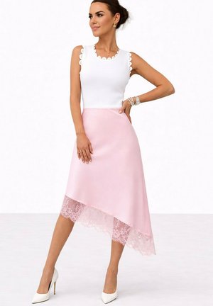 Femme posant dans un haut blanc sans manches avec des bords festonnés et une jupe asymétrique rose pâle avec une bordure en dentelle, portant des talons hauts blancs.