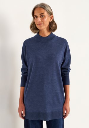 Frau mit kurzem grauem Haar, die einen locker sitzenden, langärmeligen navyblauen Pullover und dunkle Hosen trägt, steht vor einem schlichten Hintergrund.