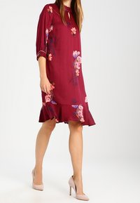 Bordeauxfarbenes Kleid mit Dreiviertelärmeln, floralem Muster, gerafftem Saum, aus leichtem Stoff, kombiniert mit hellrosa High Heels.