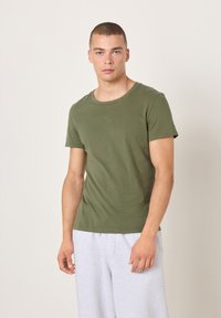 Alyvuogių žalios spalvos medvilniniai marškinėliai su apvalia iškirpte ir trumpomis rankovėmis, turintys lygią tekstūrą. Derinami su šviesiai pilkomis sportinėmis kelnėmis.