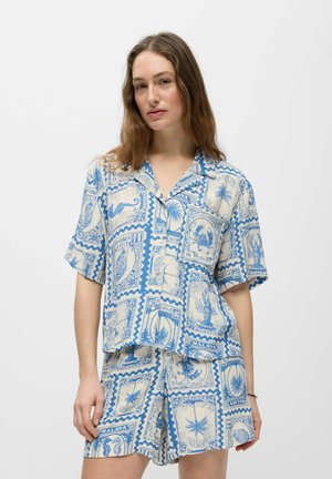 Femme portant une chemise boutonnée à manches courtes assortie bleu et blanc et un short avec des motifs de timbres-poste vintage et tropicaux.