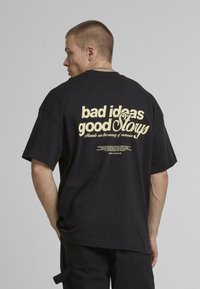 Tricou negru supradimensionat cu text imprimat de culoare crem pe spate: "idei proaste povești bune," cu text suplimentar mai mic dedesubt.