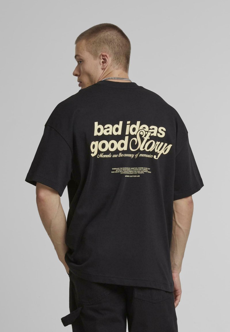 Tricou negru supradimensionat cu text imprimat de culoare crem pe spate: "idei proaste povești bune," cu text suplimentar mai mic dedesubt.
