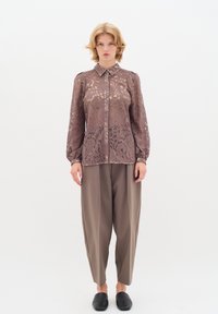 Blusa in pizzo marrone con collo e maniche lunghe, caratterizzata da intricati motivi floreali. Abbinata a pantaloni grigi larghi e mocassini neri.