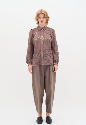Blusa in pizzo marrone con collo e maniche lunghe, caratterizzata da intricati motivi floreali. Abbinata a pantaloni grigi larghi e mocassini neri.