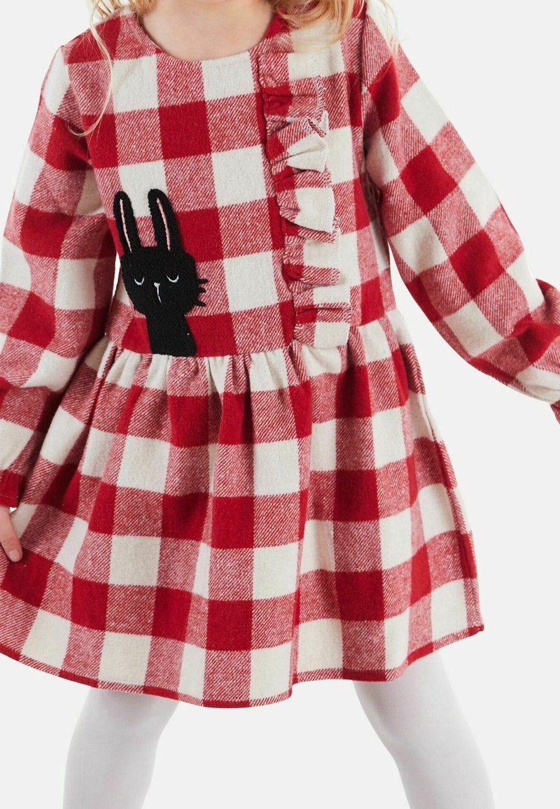 Enfant portant une robe à carreaux rouges et blancs avec manches longues, volantée à l’avant, et un détail brodé de lapin noir sur la poitrine.