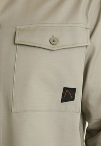 Šviesiai béžinė marškiniai su krūtinės kišenėmis ir mygtuku, tekstūruota medžiaga bei juoda pleistras su oranžiniu trikampiu dizainu.