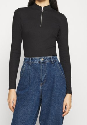 Žena nosící černý top s dlouhým rukávem, předním zipem a volné modré džínové kalhoty s vysokým pasem.