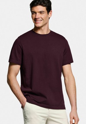 Glunderende man die een donkerpaars T-shirt met korte mouwen en ronde hals draagt en lichtbeige broek met één hand in zijn zak tegen een effen achtergrond.