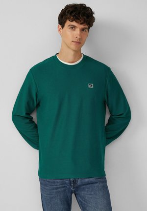 Green long-sleeve shirt featuring a round neck, ribbed texture, and a small logo on the chest. Paired with denim jeans.